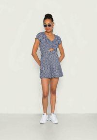 Mulher vestindo um vestido curto floral azul com recorte frontal, tênis brancos e óculos de sol azuis, de pé contra um fundo claro e liso.