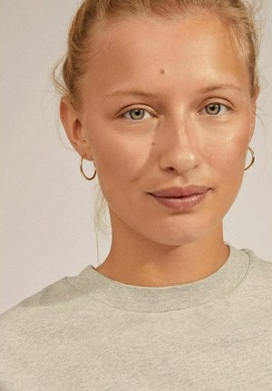 Grauer Sweatshirt mit rundem Ausschnitt; das Modell hat helles Haar und trägt kleine goldene Creolen. Der Hautton ist hell mit minimalen Unreinheiten.