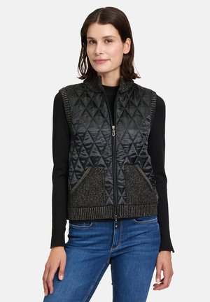 Black quilted vest with a zip front, textured collar, and beaded accents on pockets. Worn over a black long-sleeve top with blue jeans.