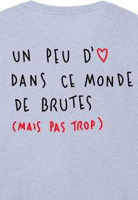Light blue shirt featuring black and red text: “Un peu d’❤️ dans ce monde de brutes (mais pas trop).” Emphasizes kindness with humor.