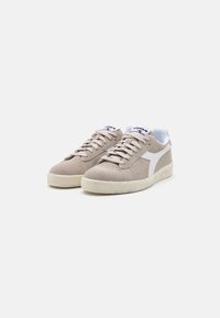 Beige suede sneakers with white accents, round toe, lace-up closure, and textured rubber sole. Features a logo on the tongue.