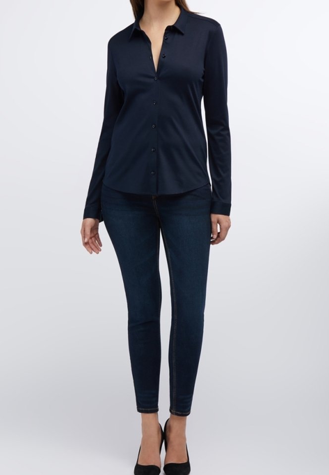 Femme portant un chemisier noir ajusté, un jean skinny foncé et des talons hauts noirs, debout contre un fond clair uni.