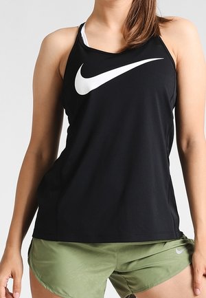 Nainen, jolla on musta hihaton Nike-toppi, jossa suuri valkoinen Swoosh-logo, ja oliivinvihreät Nike-shortsit tasaisen taustan edessä.