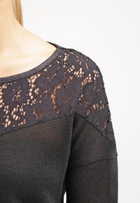 Black sweater featuring a lace overlay at the shoulders, knitted texture, and a wide neckline. Accents provide a contrasting, open pattern.