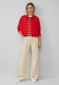 Rote Jeansjacke mit zwei Brusttaschen, kombiniert mit einem weiß-rot gestreiften Hemd und beigen weit geschnittenen Hosen. Weiße Sneaker runden das Outfit ab.