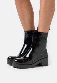 Bottines en cuir verni noir avec un talon épais, un bout rond et une semelle texturée ; elles présentent un logo circulaire sur le côté extérieur.