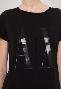 Černé tričko s krátkým rukávem s lesklým, vyvýšeným logem „AX“ a názvem značky „ARMANI EXCHANGE“ v elegantním písmu.