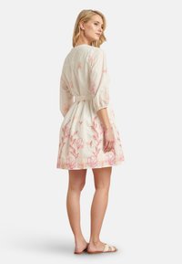 White dress with pink floral embroidery, three-quarter sleeves, cinched waist, and flared skirt. Worn with flat, beige sandals.