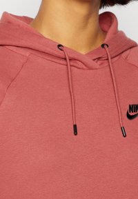 Rosa hoodie med dragsko, prydd med en svart Nike-logotyp. Tillverkad av mjukt tyg med en slät yta och raglanärmar.