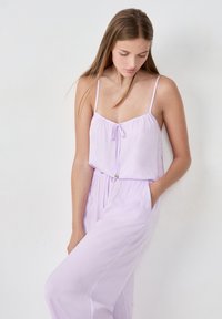 Liliowy kombinezon na cienkich ramiączkach, z regulowanym w pasie i luźnym krojem. Wykonany z lekkiego materiału o gładkiej fakturze.