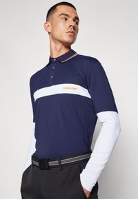 Ναυτική polo μπλούζα με λευκή οριζόντια γραμμή και πορτοκαλί λογότυπο; διαθέτει γιακά, τρία κουμπιά και κοντά μανίκια πάνω από μία μακριά λευκή μπλούζα.