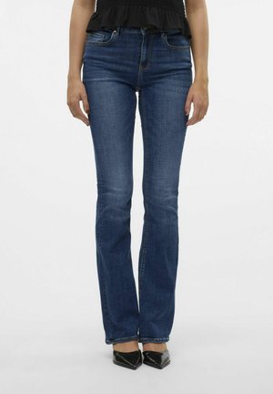 Kobieta w dopasowanych niebieskich jeansach typu bootcut, czarnych szpiczastych butach na obcasie oraz czarnym topie, stojąca na jednolitym białym tle.