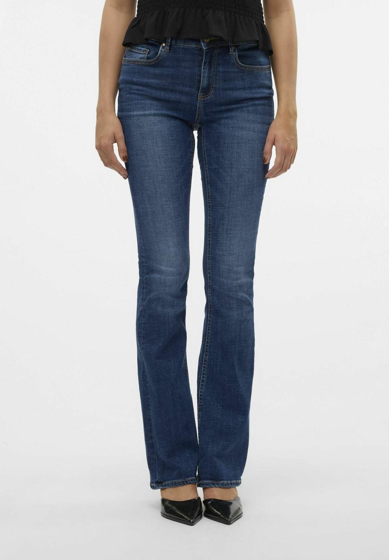 Femme portant un jean bootcut bleu ajusté, des chaussures noires à talons pointues et un haut noir, debout devant un fond blanc uni.