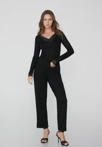 Schwarzer Langarm-Bodysuit mit Spitzen-Halsausschnitt, kombiniert mit maßgeschneiderten schwarzen Hosen und schwarzen Riemchen-Absätzen. Glattes Material und taillierte Passform.