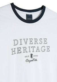 Biała koszulka z granatowym prążkowanym dekoltem. Zawiera nadrukowany tekst: "DIVERSE HERITAGE EST. 1993" na czarno, wraz z logo i detalem poniżej.