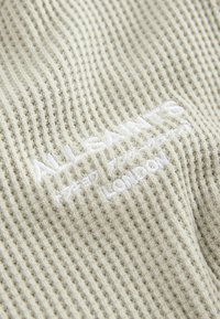 Tessuto grigio chiaro con texture a waffle e branding ricamato bianco. Presenta una superficie morbida e rigata con una leggera lucentezza.