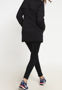 Svart puffjacka med hög krage, långa ärmar och insvängd midja. Bärs tillsammans med svarta leggings och marinblå träningsskor.