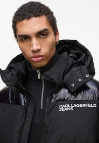 Zwarte gewatteerde jas met glanzende accenten, ritssluiting en een capuchon. Voorzien van het logo "KARL LAGERFELD JEANS" in het wit op de borst.