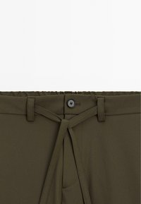 Pantalons vert olive avec une fermeture à bouton, des passants de ceinture, et une ceinture en tissu assorti nouée à l'avant de la taille.