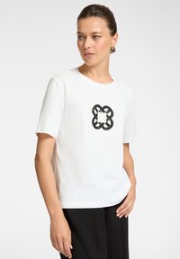 Witte katoenen t-shirt met een zwart geborduurd knoopontwerp in het midden. Korte mouwen en een losse pasvorm, gecombineerd met zwarte broek.