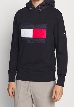 Man in een zwarte hoodie met een grote, gequiltte rechthoekige patch in rood, wit en blauw op de borst en beige broek.