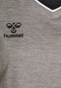 Primo piano di una maglia sportiva grigia in tessuto testurizzato con colletto a V nero e bianco e logo "hummel" nero con emblema stilizzato di ape.