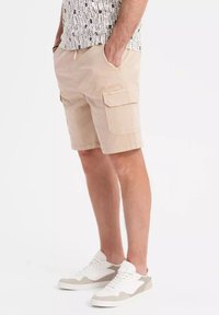 Shorts cargo beiges avec plusieurs poches, une coupe décontractée et une taille élastique à cordon, associés à des sneakers blancs et gris.