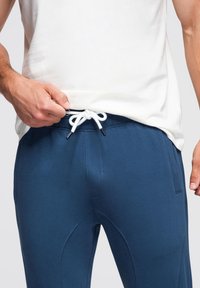 Blauwe sportbroek met een tailleband met trekkoord, voorzien van een witte koord en metalen uiteinden. Glad materiaal met zijzakken en een getailleerd ontwerp.
