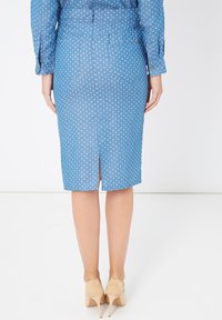 Mujer con una falda lápiz azul con lunares y camisa a juego, zapatos de tacón bajos beige, de pie con las manos relajadas a los lados.