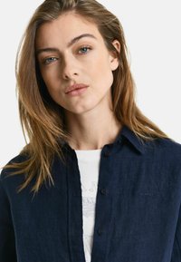 Navyblauw linnen overhemd met knoopsluiting en een kraag, met subtiele branding op de borst en gedragen over een wit graphic T-shirt.