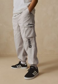 Pantaloni cargo gri deschis cu manșete elastice, numeroase buzunare și accente negre. Asortați cu adidași negri cu dungi albe.