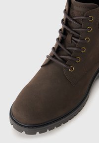 Botín de ante marrón con ojales de metal, cordones marrones y una suela de tracción resistente con punta redondeada. Textura suave y diseño robusto.