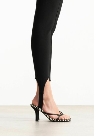 Close-up van een voet met zwarte, gestripte zebra-print sandalen met hoge hakken en bandjes, gecombineerd met zwarte nauwsluitende broek met een voorste stijgbeugelontwerp.