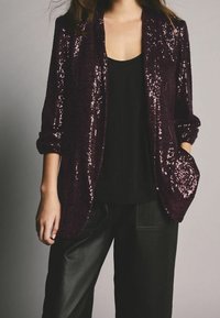 Bordeaux paljet-blazer med tekstureret overflade, med en enkelt lomme, parret med en sort tanktop og læderbukser.