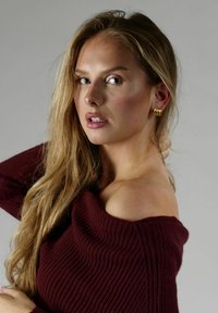 Burgunderribs strikket sweater med off-shoulder design, parret med guld hoops smykker. Lang, lige blond hår. Neutral baggrund.