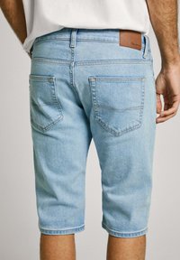 Pantalones cortos de mezclilla azul claro con corte recto, que presentan dos bolsillos traseros y un parche de cuero en la cintura. Textura suave y ajuste entallado.