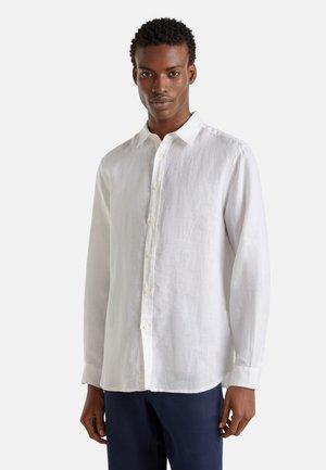 Biała lniana koszula z długim rękawem, z kołnierzykiem zapinanym na guziki, plisą z przodu oraz mankietami. Lekko teksturowany materiał, luźny krój, noszona z granatowymi spodniami.