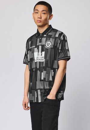Man draagt een poloshirt met zwart- en grijs geometrisch patroon en het logo en de tekst "Weekend Offender", staand met één hand in de zak.