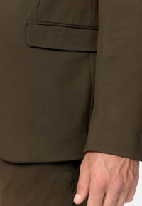 Giacca marrone con revers a punta, dotata di una tasca sul petto e una texture del tessuto liscia. Primo piano della manica e della mano.
