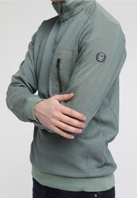 Zielona bluza z zamkiem na szyi, z teksturowanymi wstawkami, kieszenią na piersi i akcentem z logo na rękawie. Dopasowane mankiety i dół dopełniają design.
