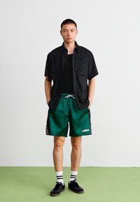Černá košile na knoflíky, zelené sportovní šortky s bílými pruhy, černé tenisky a pruhované ponožky. Jednoduchý design s detailem kapsy.