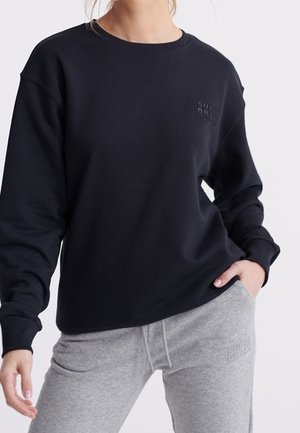 Person iført en sort sweatshirt og grå joggingbukser, med den ene hånd i lommen, beskåret fra nakken til midt på låret på en ensfarvet baggrund.
