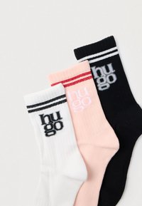 Trīs pāri ribotiem zeķu pāriem baltā, rozā un melnā krāsā, ar kontrastējošām svītrainām aprocēm un "hugo" logotipu treknā drukā.