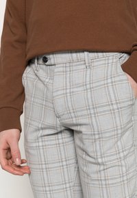 Grå rutiga byxor har en blandning av grå, svart och bruna linjer. Tyget har en mjuk textur med en knappknäppning fram och sidofickor.