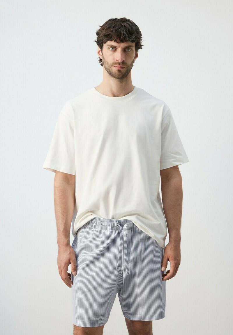 Mann trägt ein schlichtes weißes, übergroßes T-Shirt und blau-weiß gestreifte, mit Kordelzug versehene Shorts mit vertikalen Streifen und steht vor einem schlichten Hintergrund.