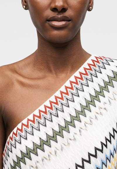 Κοντινό ενός ατόμου που φοράει μπλουζάκι με έναν ώμο με κόκκινα, μαύρα, πράσινα και μπλε ζιγκ-ζαγκ μοτίβα και μικρά χρυσά καρφωτά σκουλαρίκια.