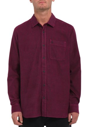 Homme portant une chemise à manches longues à boutons avec un petit motif à carreaux bordeaux et noir, dotée d'une poche poitrine.