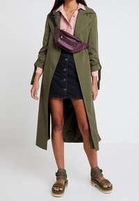 Lungo trench coat verde oliva indossato sopra una camicia button-up rosa chiaro, una gonna di jeans nera e sandali piatti verdi. È indossata una borsa a tracolla color marrone bordeaux.