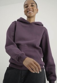 Fialová mikina s kapucí a klokankovým kapsou, žebrovanými manžetami a hladkým materiálem. Nosená s tmavými džíny.