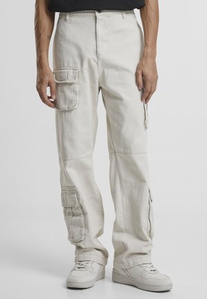 Pantaloni cargo bej cu multiple buzunare laterale, cu croială dreaptă și din material de bumbac. Purtați cu adidași albi.